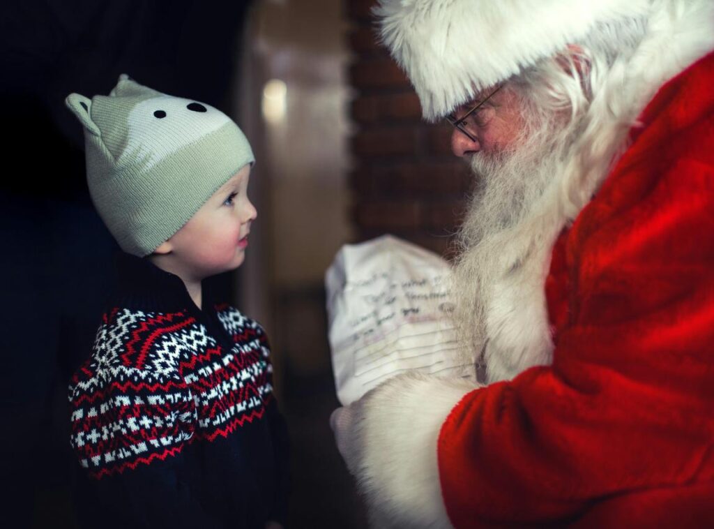A child in a bear hat talks to Santa, who holds a letter. Both are engaged in conversation.