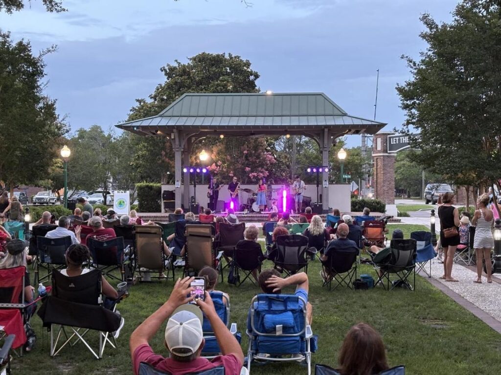 A crowd enjoys a live band performance on a stage in a park during twilight.