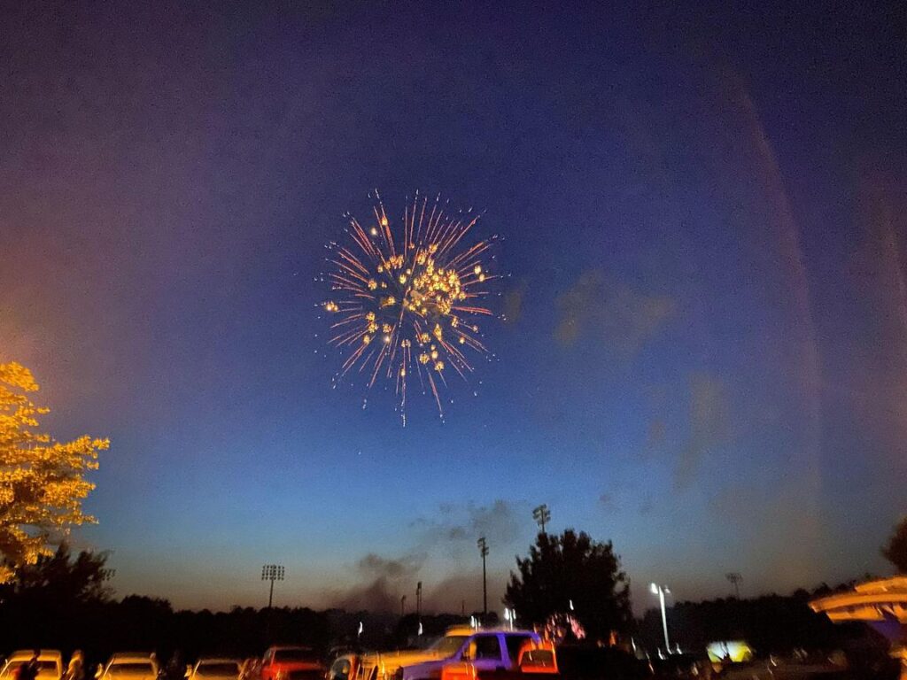 Fireworks burst in the night sky above a parking lot, with trees and stadium lights in the background.