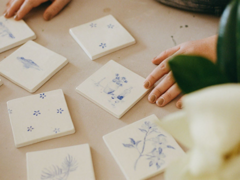 Hands arrange decorative blue and white tiles on a table, surrounded by a plant.
