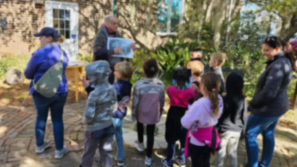 An adult shows a picture to a group of children outdoors, surrounded by greenery.