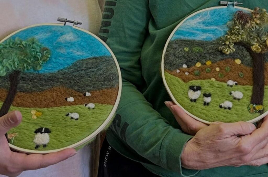 Two people hold embroidered hoop art featuring sheep and a landscape with trees and hills.