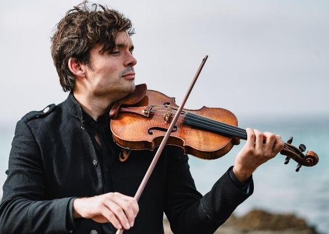 A man plays the violin outdoors, focused on his music, with the ocean in the background.