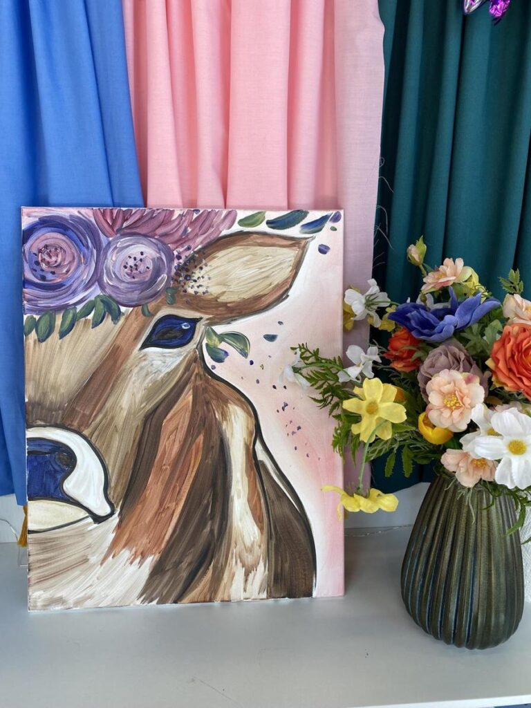 A painted cow portrait with flowers beside a vase, set against colorful fabric backdrops.