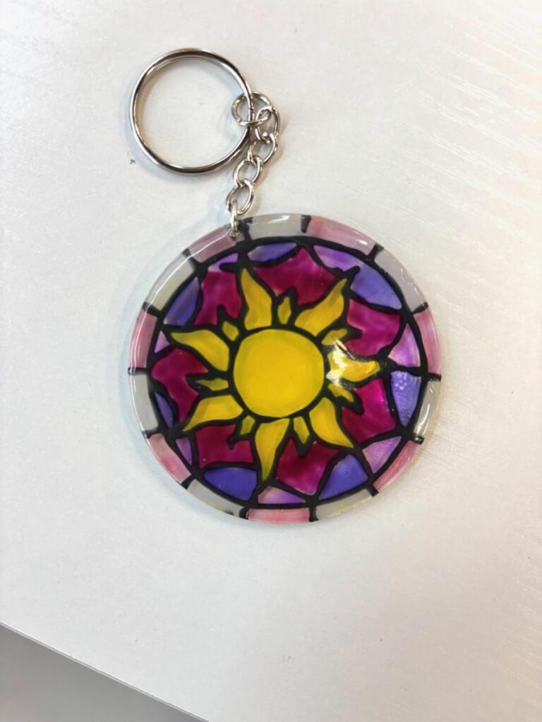 A colorful keychain featuring a sun design surrounded by purple and pink stained glass patterns.