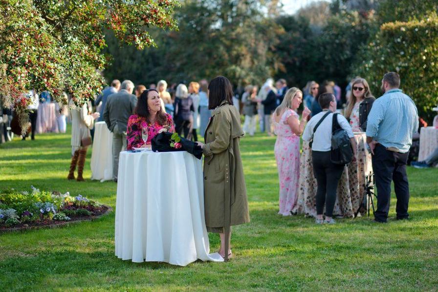 Two women converse at a table in a garden, while others socialize in the background.