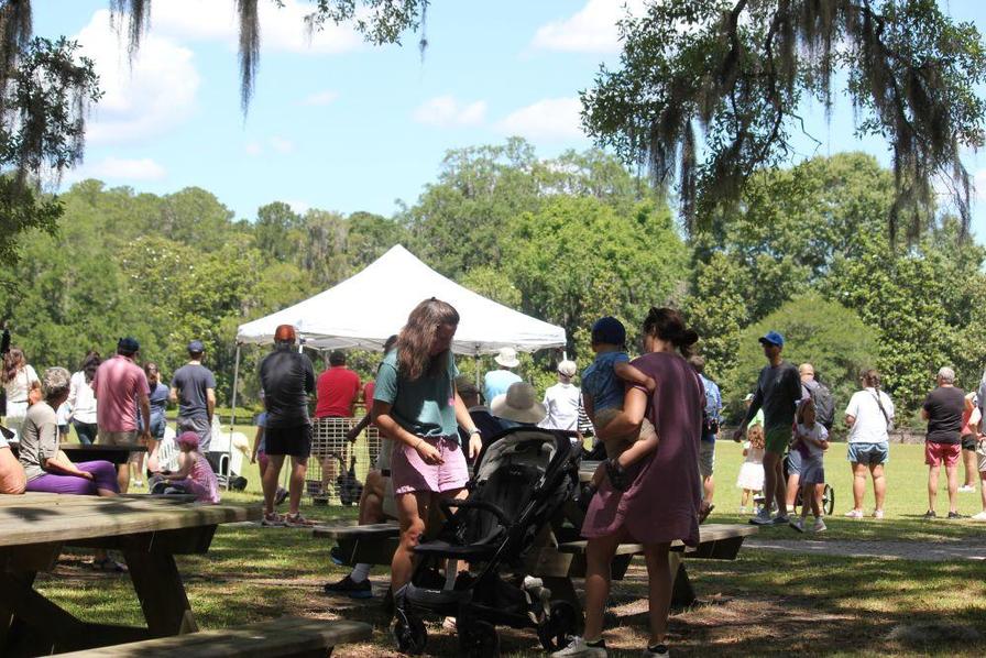 People gather at a park event, with a tent in the background and children playing nearby.
