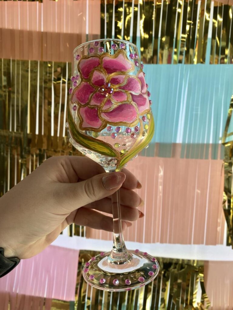 A hand holds a decorative glass with a pink flower design and colorful gems, against a shiny backdrop.