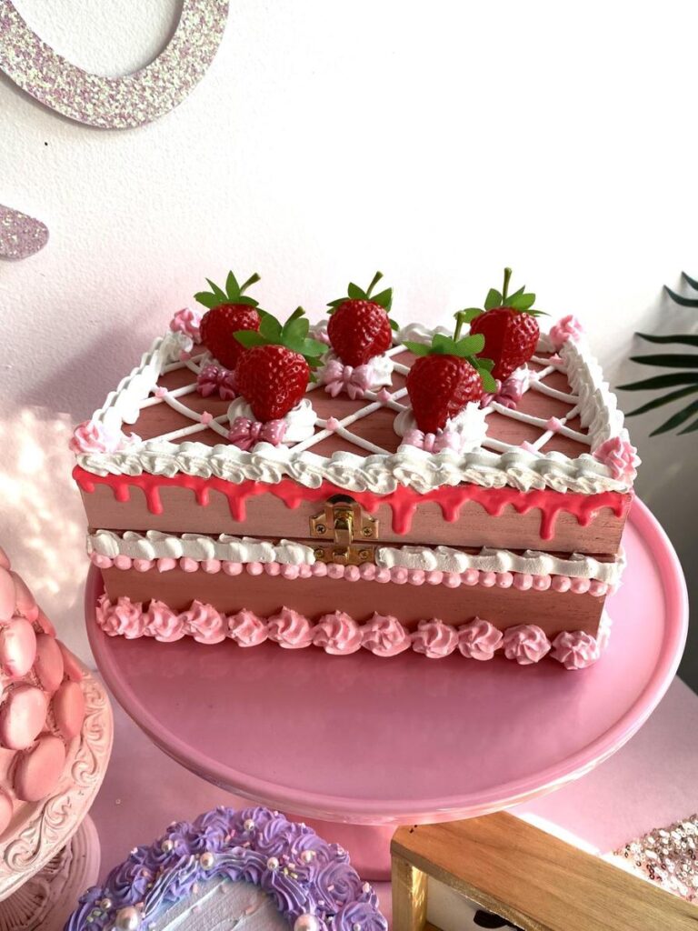 A decorative cake with strawberries and icing sits on a pink cake stand, surrounded by other desserts.