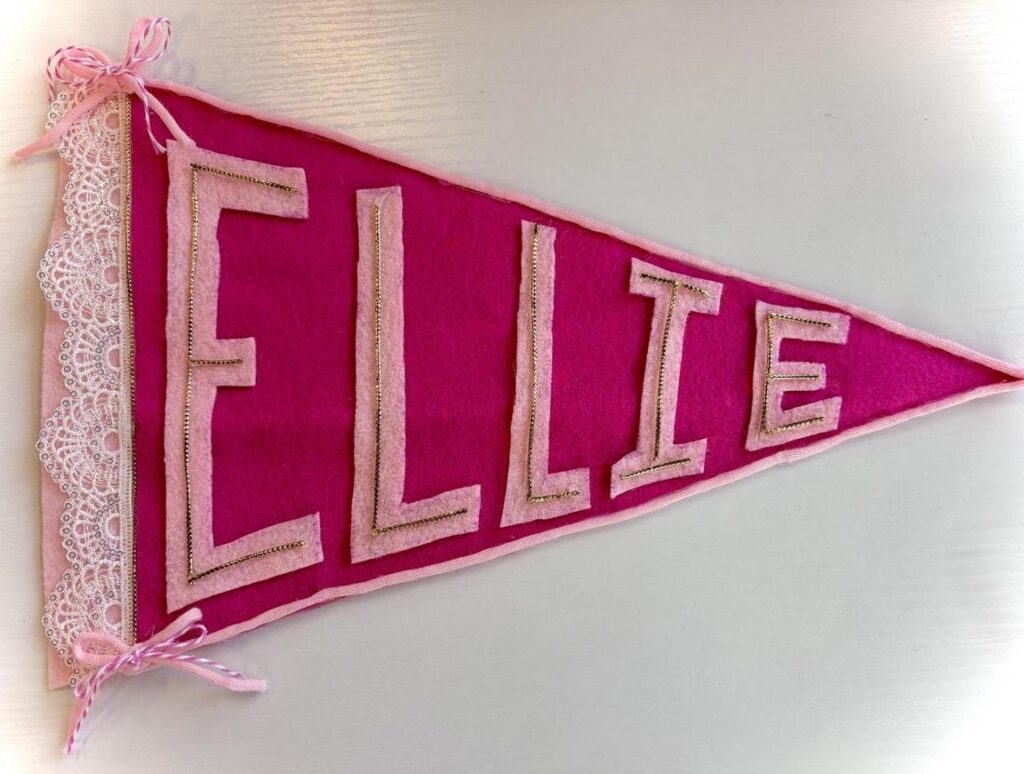 Pink felt pennant with the name "ELLIE" in gold and pink letters, decorated with lace and ribbon.