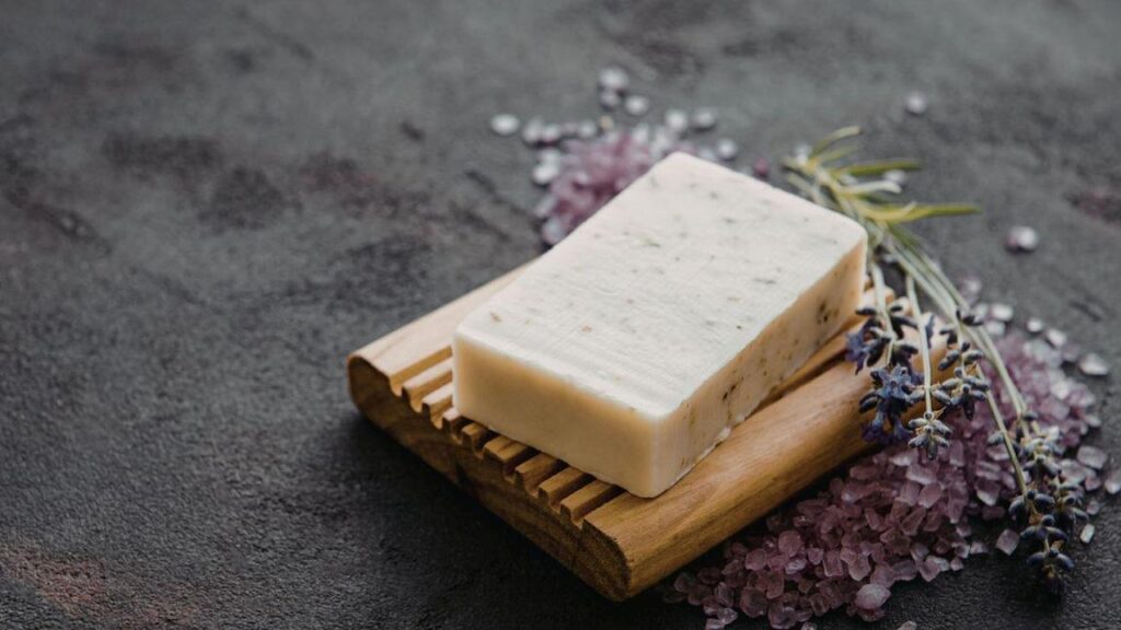A bar of soap with herbs rests on a wooden dish, surrounded by pink salt and lavender sprigs.