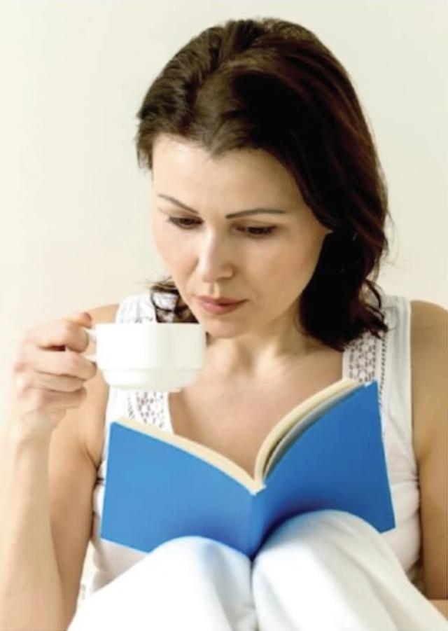A woman sits comfortably, reading a blue book while holding a cup.