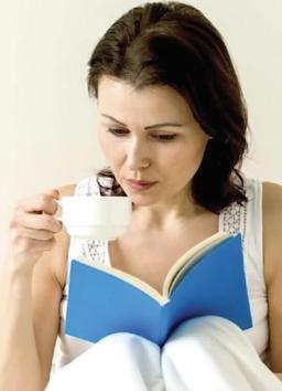 A woman sits comfortably, reading a blue book while sipping from a white cup.