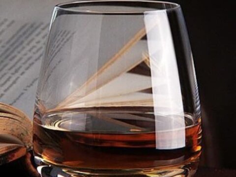A clear glass filled with amber liquid sits on a wooden surface, with blurred text in the background.