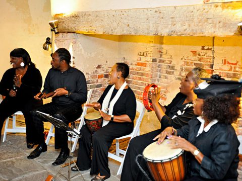 Five musicians perform together, playing drums and tambourine, in a cozy, rustic setting.