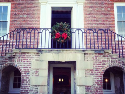 A festive wreath with red ribbons hangs on a balcony above a stone entrance. Brick walls frame the scene.