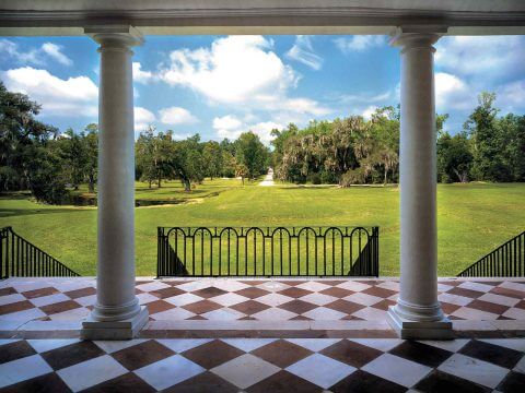 View from a portico with columns overlooking a green landscape and trees under a blue sky.