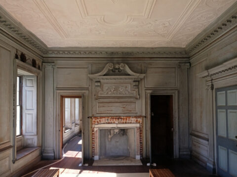 Interior of a room with ornate ceiling, fireplace, and two doorways. Light enters through windows.
