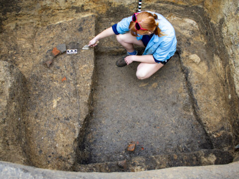 A person with red hair kneels in an archaeological dig site, using a brush to uncover artifacts.